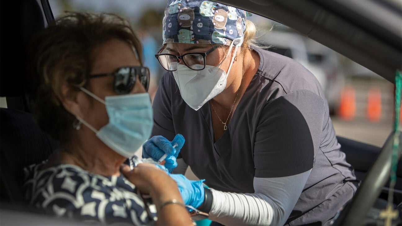 A healthcare worker in an N95 mask administers a vaccine to a person sitting inside their car