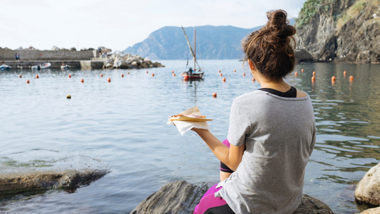 A woman eats a slice of pizza while sitting on rocks along a shoreline