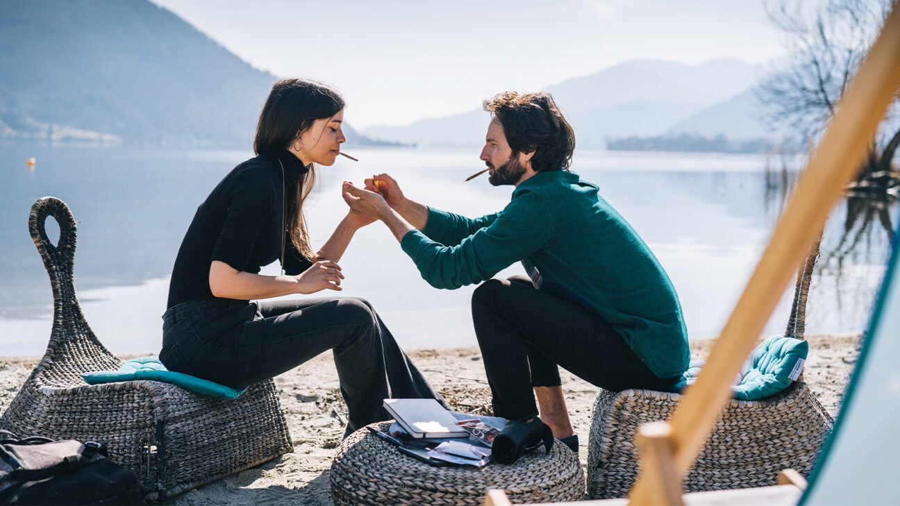 A young man and young woman light up cannabis cigarettes while sitting along a lake