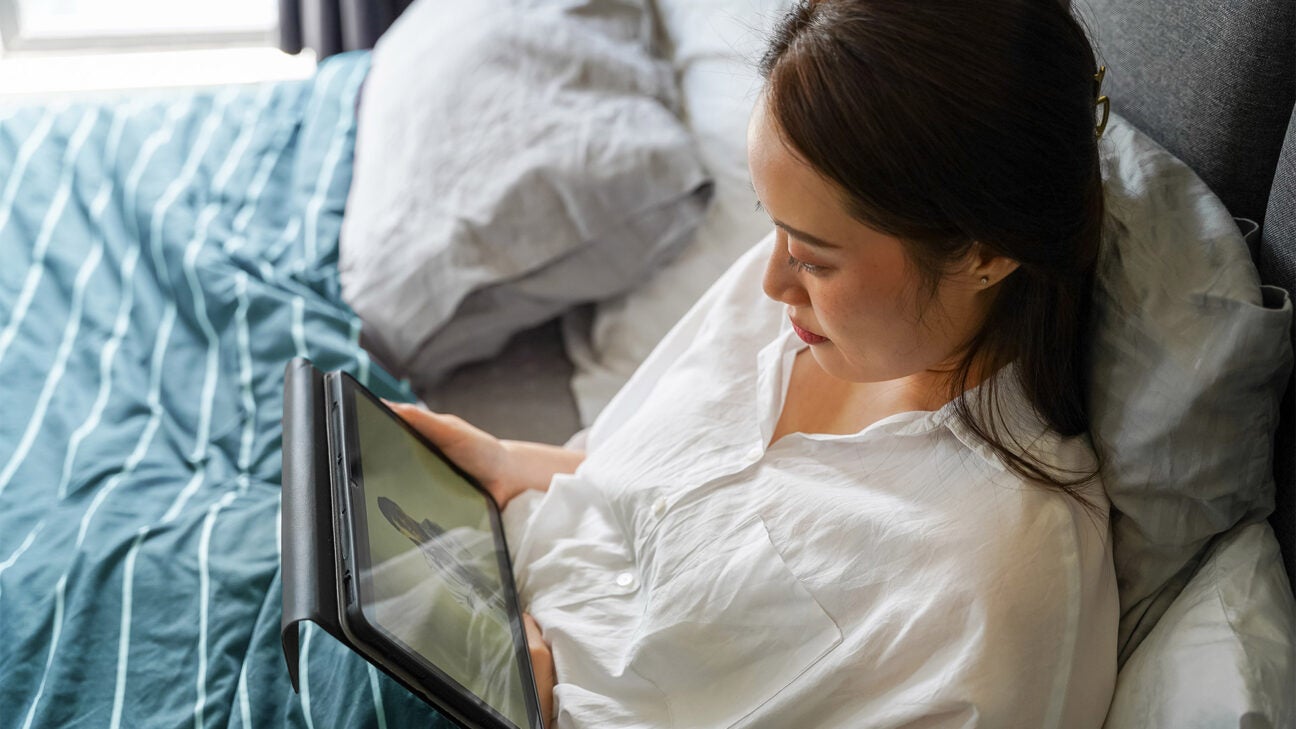 A young woman rests in bed while conducting a telehealth meeting via an iPad