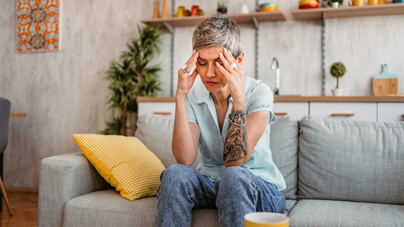 A woman closes her eyes and holds her head while sitting on a couch