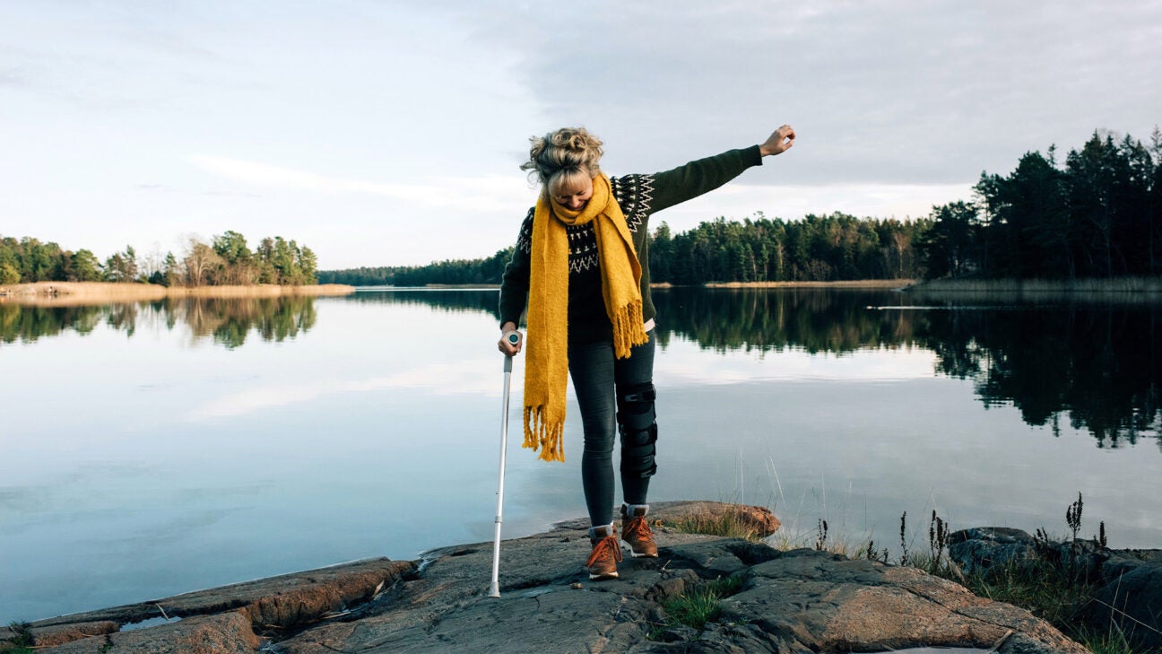 A woman with a cane walks on some rocks along a lake