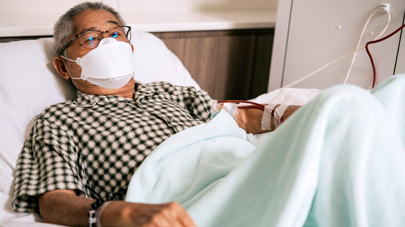 An older man wearing a mask rests in a hospital bed