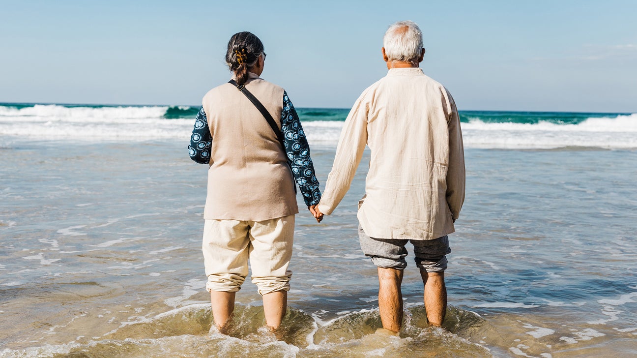 An older woman and man hold hands while standing in the shallow waters of an ocean