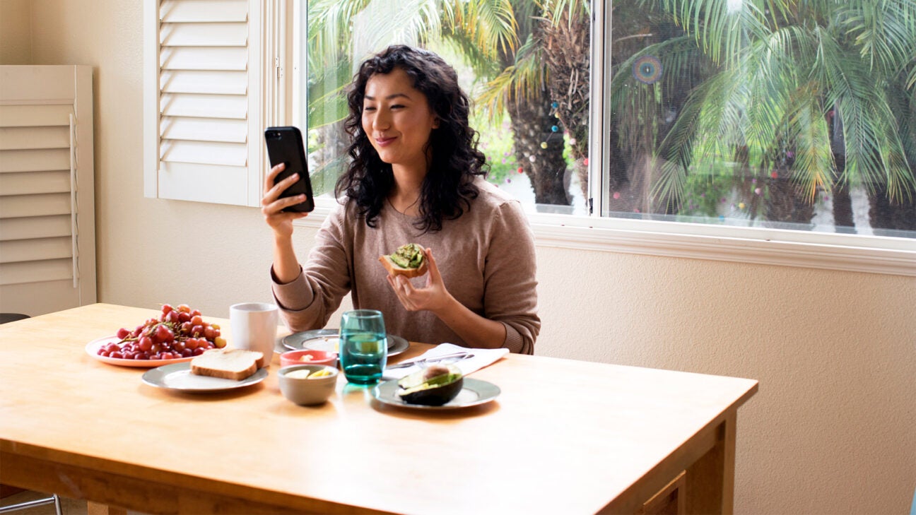 A woman smiles while eating fruit and checking an app on her phone in her dining alcove