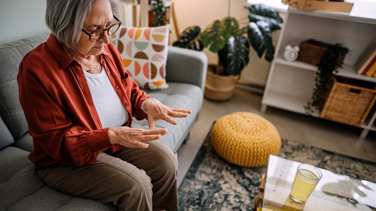 An older woman studies her hands while sitting on a couch in her living room