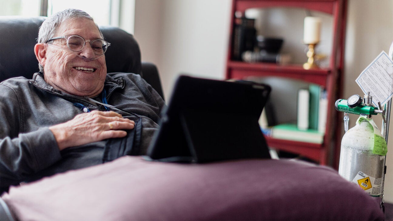 A man with COPD smiles while looking at a laptop while resting in bed