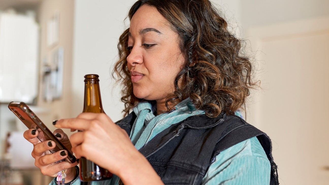 A woman looks at her phone while holding a beer.