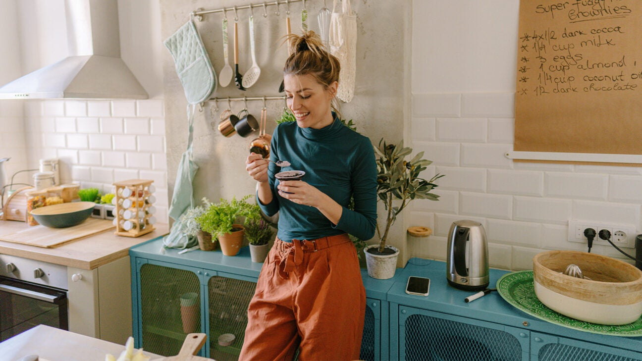 A woman standing in a kitchen eating yogurt.