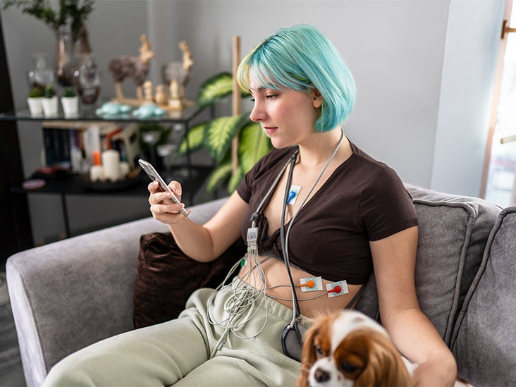 A woman wearing a heart monitor sits on a sofa next to a dog.